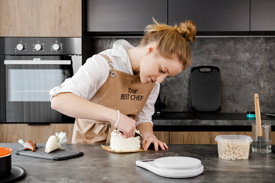 A young, smiling pastry chef in a beige apron prepares a small cake with a offset spatula in a modern kitchen. Close up.
