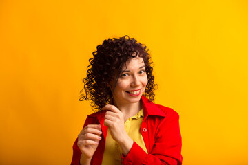 Studio shot of positive and smiling pretty female. Curly girl touching her hair. Young woman in bright red shirt. Isolated on yellow background. Close up.