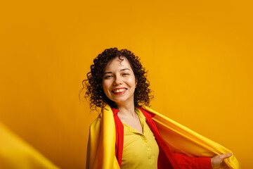 Studio shot of positive young woman. Cute smiling female wraps herself in yellow cloth. Curly girl in bright red shirt and yellow cloth. Isolated on yellow background.