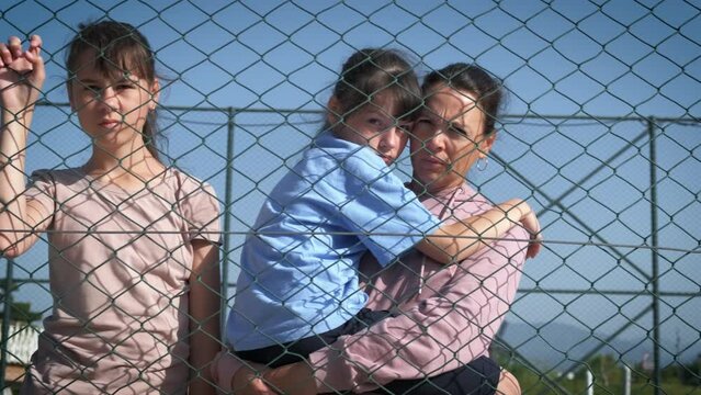 Family in a refugee camp. A desperate woman with a child in her arms and a teenage daughter looks sadly through the iron mesh. Refugees in a refugee camp.