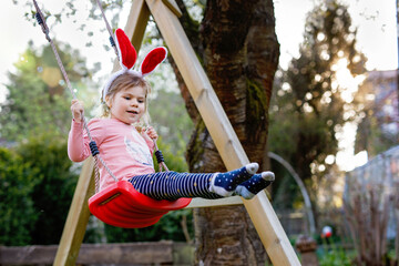 Happy beautiful little toddler girl with red Easter bunny ears having fun on swing in domestic...