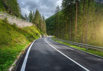 Road in green forest at sunset in summer. Dolomites, Italy. Beautiful mountain roadway, tress, grass, high rocks, blue sky with clouds. Landscape with empty highway through the wood in spring. Travel - obrazy, fototapety, plakaty