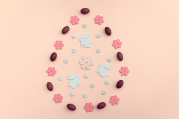 Easter flat lay with white wooden bunny cutouts and pink flowers, chocolate eggs,  arranged in an egg shape on pink background. A festive spring concept for holiday designs and decorations.