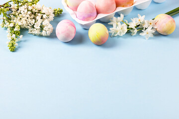 Colorful Easter eggs in a white stand with spring blooming daffodils and white lilac on a blue background. Happy Easter concept. Greeting card, copy space