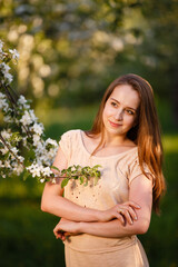 Young woman in beige dress in a blooming apple garden. Spring story. Brown-haired romantic girl with long hairs. Woman plays with her hairs. Woman portrait. Walk in a spring park