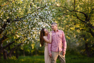 Young man and woman couple in a blooming apple garden. Tender holding each other. Spring lovestory. Brown-haired girl with long hairs and man in pink shirt. Young family. Couple romantic spring date