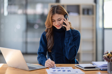 Attractive successful young business Asian woman in striped blouse working in modern office, making phone call to potential client, having nice conversation, sitting at desk in front of open laptop