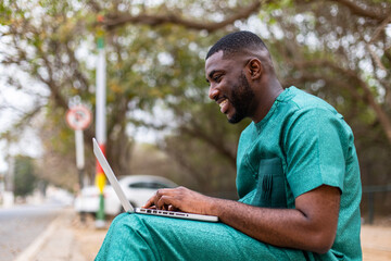 Young Millennial African male typing and smiling on laptop computer outdoors, addicted to technology and social media.