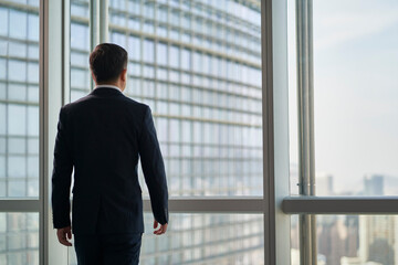 Fototapeta premium rear view of an asian businessman walking up to windows in office looking at city view