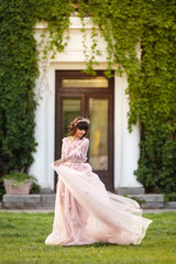 Fototapeta premium Beautiful woman in flying airy pink dress on background of door overgrown with wild grapes. Pretty young caucasian girl plays with her dress. Girl dance in flowing dress