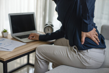 Young man sitting at the desk. He used hand holding to press down on the lower back. He felt unhappy due to pain in the lower back area due to long hours of sitting and office syndrome. - obrazy, fototapety, plakaty