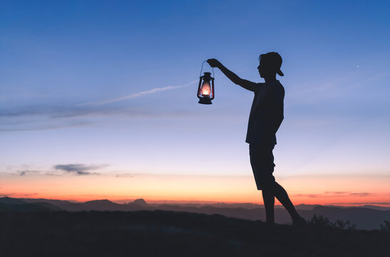 Silhouette of a man holding a lit kerosene lamp to light his way in the dark, with sky after sunset in the background. Concept of faith and hope in the face of adversity.