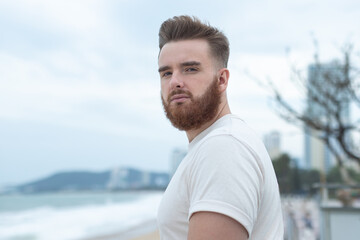 Fototapeta premium Portrait of young handsome bearded pensive man with beard is enjoy walk on summer beach near sea, breathe deep fresh air in tropical country. Summer vacation.