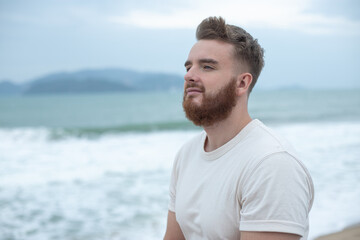 Portrait of young handsome bearded pensive man with beard is enjoy walk on summer beach near sea,...