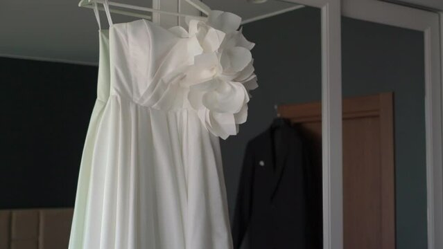 A beautiful white wedding dress hangs on a hanger on the wardrobe in the bedroom. Morning of the bride, wedding day. Women's fashionable clothes for the holiday. Black groomjacket.