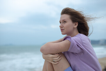 Young calm sad serious woman is sitting on embankment near sea, ocean on beach, girl thinking,...