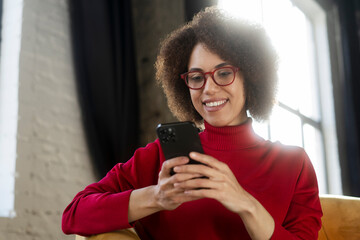 Beautiful smiling African American woman holding mobile phone watching videos sitting at home. Modern female wearing red eyeglasses using mobile app, shopping online, ordering food. Mobile banking