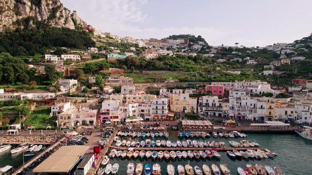 An ascending aerial shot from the boats in busy Marina Grande, over stunning cliffs and winding roads up to the boutique shops, fashionable caf&eacute;s and luxury hotels of Capri Town in Campania, Italy.