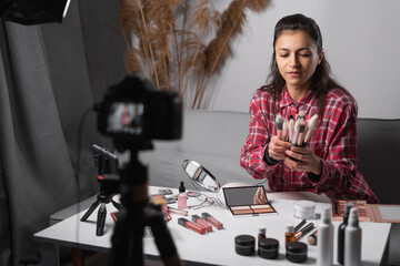 Beauty blogger or vlogger filming daily make-up routine tutorial on camera, showing make up brushes. Influencer girl livestreaming cosmetics product review in home studio.