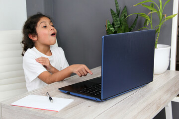 Fototapeta premium 9-year-old Latino girl does home schooling takes online classes at home on a desk with a laptop, studies, is surprised and participates in class