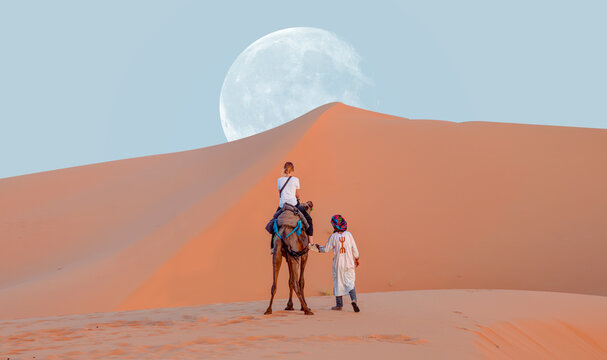 A woman riding a camel across the thin sand dunes of the in Western Sahara Desert with full moon - Morocco, Africa