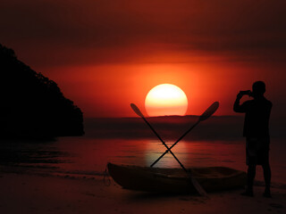 Silhouette of asian man stand and take sunset photo on beach near kayak in evening,