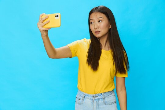 Asian woman holding her phone and looking at the screen taking pictures of herself talking on a video call with a yellow case on a blue background wearing a yellow T-shirt smiling with teeth 
