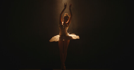 Young woman ballerina in white tutu, dancing on pointe with arms overhead, in the studio against a...