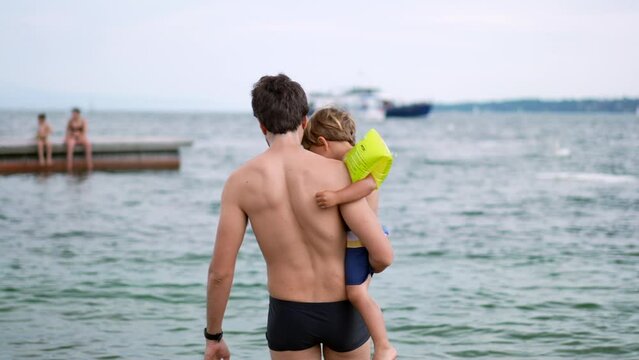 Back Of Father Holding Son In Arms Entering Water At Lake Or Beach. Child Wearing Arm Floaties Sleeves. Fatherhood Lifestyle Concept