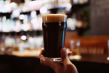 glass of dark stout beer with foam and bubbles in hand of a man in pub behind the bar - obrazy, fototapety, plakaty