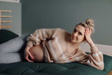 Attractive pregnant woman in pink sweater lies relaxed on sofa, leaning on her elbow, and gently hugs her stomach. 