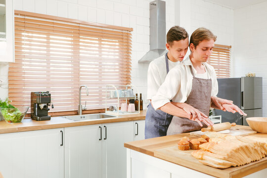 Happy Caucasian Gay Couple Making Bread Together One Person Mixes The Flour In Front. And Another Person Watching From Behind In The Kitchen At Home. LGBT Relationships. Gay Couple Concept