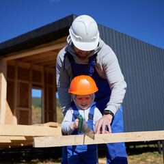 Father with toddler son building wooden frame house. Male builders using hand saw to cut boards on...