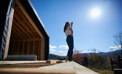 Happy woman on construction site standing on terrace at unfinished wooden frame house on sunny day.