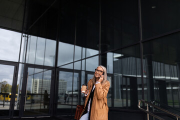 Fototapeta premium confident mature adult female accountant with gray hair speaks on the phone against the backdrop of an office building