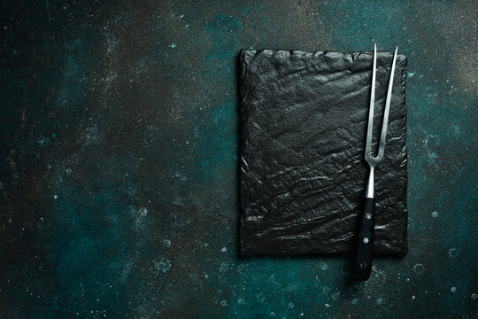 Slate plate and Metal fork for meat and steaks. On a dark green-turquoise background. Free space for text.