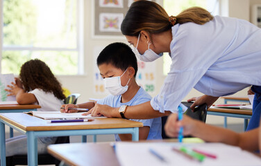 Obraz na płótnie Canvas Education, covid and learning with face mask on boy doing school work in classroom, teacher helping student while writing in class. Elementary child wearing protection to stop the spread of a virus