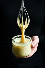 Fresh homemade sweetened condensed milk in a jar and a kitchen whisk. On a black background.