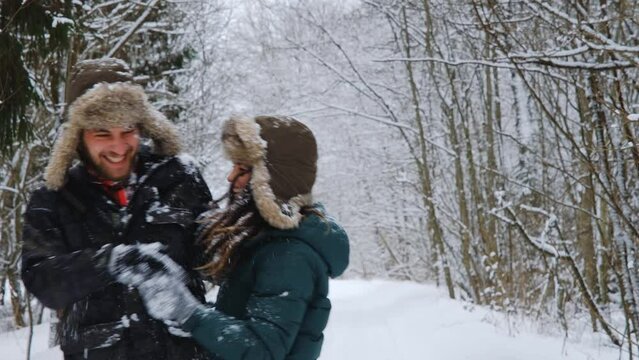 Lifestyle concept relationship between man woman in love. Young European couple playing snowballs in forest, rejoicing and laughing. Guy with girlfriend in earflaps during snowfall hugging in park.