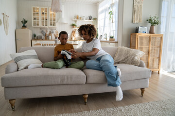 New technologies and children. Confused little African American boy experiencing virtual reality for first time while sitting on sofa with mom at home. Modern young mother giving kid son VR headset