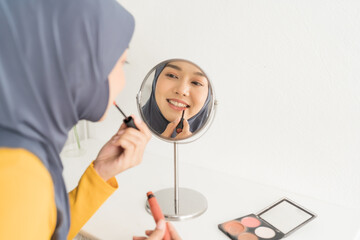 Naklejka premium Asian young Muslim woman wearing hijab headscarf applying lipstick on lips and looking at mirror at home