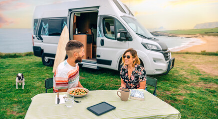 Happy young couple sitting outdoors in summer with camper van and beach on background