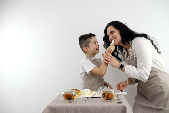breakfast mom and son try cheese son feeds mom a piece of cheese boy puts honey on a piece of cheese black tea nearby on table laughter joy pleasant pastime family smile kitchen apron
