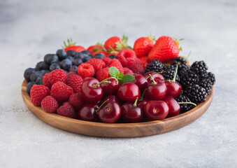 Fresh organic summer berries mix in round wooden tray on light kitchen table background. Raspberries, strawberries, blueberries, blackberries and cherries.