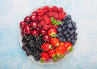 Fresh organic summer berries mix in white plate on blue kitchen table background. Raspberries, strawberries, blueberries, blackberries and cherries.