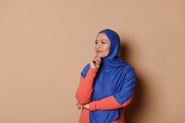 Pensive doubtful young Muslim charming woman in elegant orange dress and blue hijab, thoughtfully...