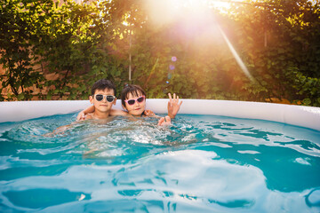 Happy children are relaxing in big swimming pool in garden outside at sunset. Wonderful sunlight greenery background.