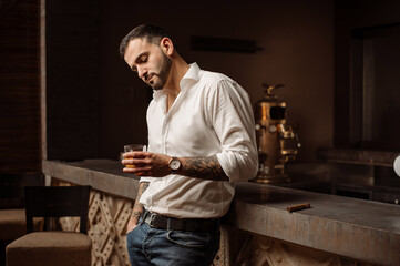 Handsome bearded male in shirt and jeans with glass of whiskey and cigar in the bar or pub