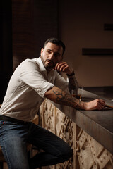 Obraz premium Handsome bearded male in shirt and jeans with glass of whiskey and cigar in the bar or pub