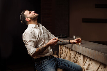 Handsome bearded male in shirt and jeans with glass of whiskey and cigar in the bar or pub